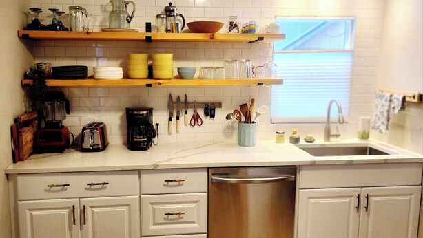 A kitchen with white cabinets, a dishwasher, a coffee maker, and various utensils on the countertop. - The Cozy Nook- Adorable 2 Bedroom Home (Columbia Falls)