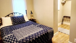 A bedroom with a bed, a nightstand, a lamp, a window with curtains, and a hallway leading to a bathroom. - The Cozy Nook- Adorable 2 Bedroom Home (Columbia Falls)