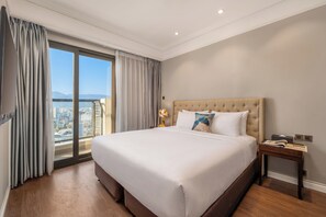 A hotel room with a large bed, a nightstand, a lamp, and a view of the city through a large window. - Luxury Apartment Seaside (Da Nang)