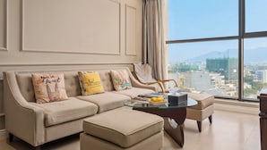 Ein modernes Wohnzimmer mit Sofa, Hocker und Glastisch, ausgestattet mit dekorativen Kissen und Blick auf eine Stadtkulisse durch große Fenster.