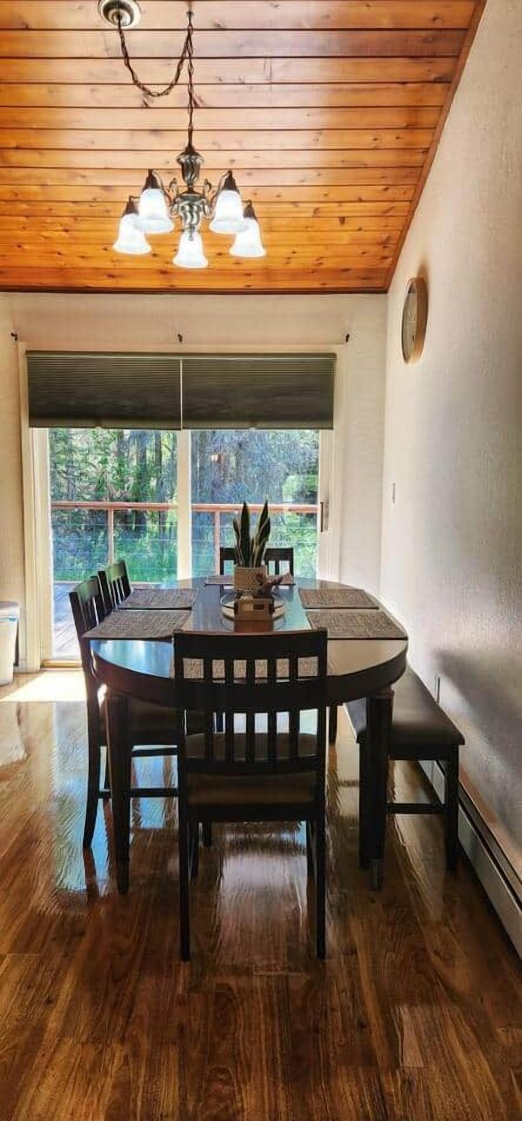 Uma sala de jantar com mesa de madeira, cadeiras, um candelabro e vista através de uma janela ampla.