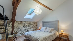A bedroom with a stone wall, a sloped ceiling, a skylight, a bed with a headboard, a bedside table with a lamp, and a wooden chair. - Domaine Le Pehau 4 - Avec jardin et terrasse (Osserain-Rivareyte)