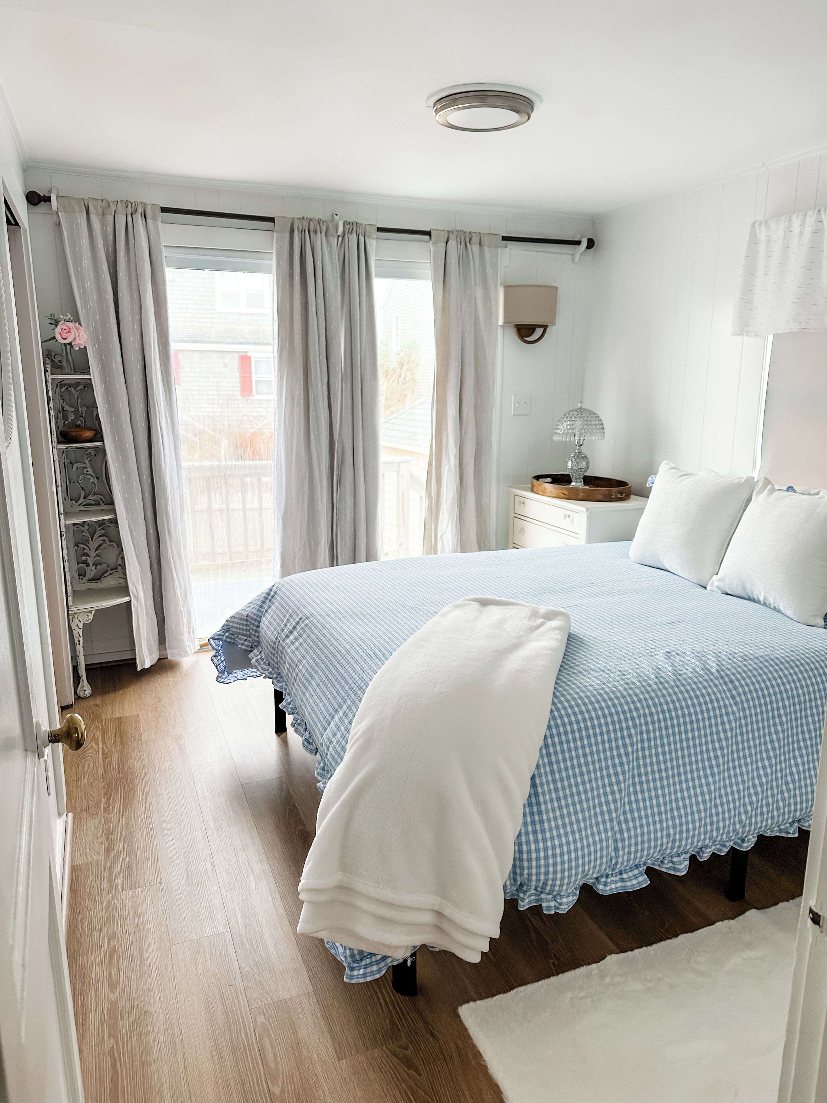 Een slaapkamer met een bed, nachtkastje, lamp, raam met gordijnen, deur en een houten vloer.