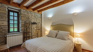 Une chambre avec un mur en pierre, des poutres apparentes, un lit avec une literie à carreaux, un radiateur et une fenêtre donnant sur un paysage verdoyant.