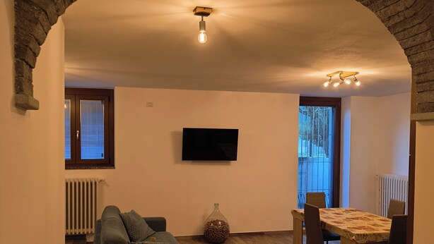 A living room with a stone archway, a sofa, a dining table with chairs, a TV on the wall, and a radiator. - Dwelling Euthymia a few minutes from Lake Como (Cosio Valtellino nr. 51)