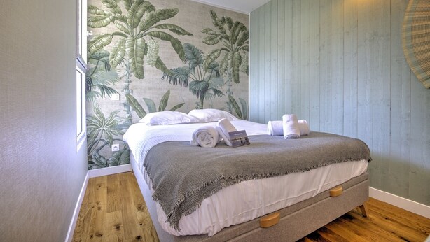 A neatly made bed with a white comforter and a gray blanket, a small bedside table with a book, and a wall with a tropical leaf pattern. - Brousse & Brise - Studio à deux pas de la mer (Arcachon)