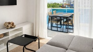 Habitació d'hotel moderna amb un sofà gran, una televisió de pantalla plana, una taula de menjador amb caires i vistes a una piscina i edificis.
