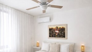 Un dormitori amb un llit, un ventilador de sostre, aire condicionat, un quadre penjat a la paret, làmpades de nit i una finestra amb cortines transparents.