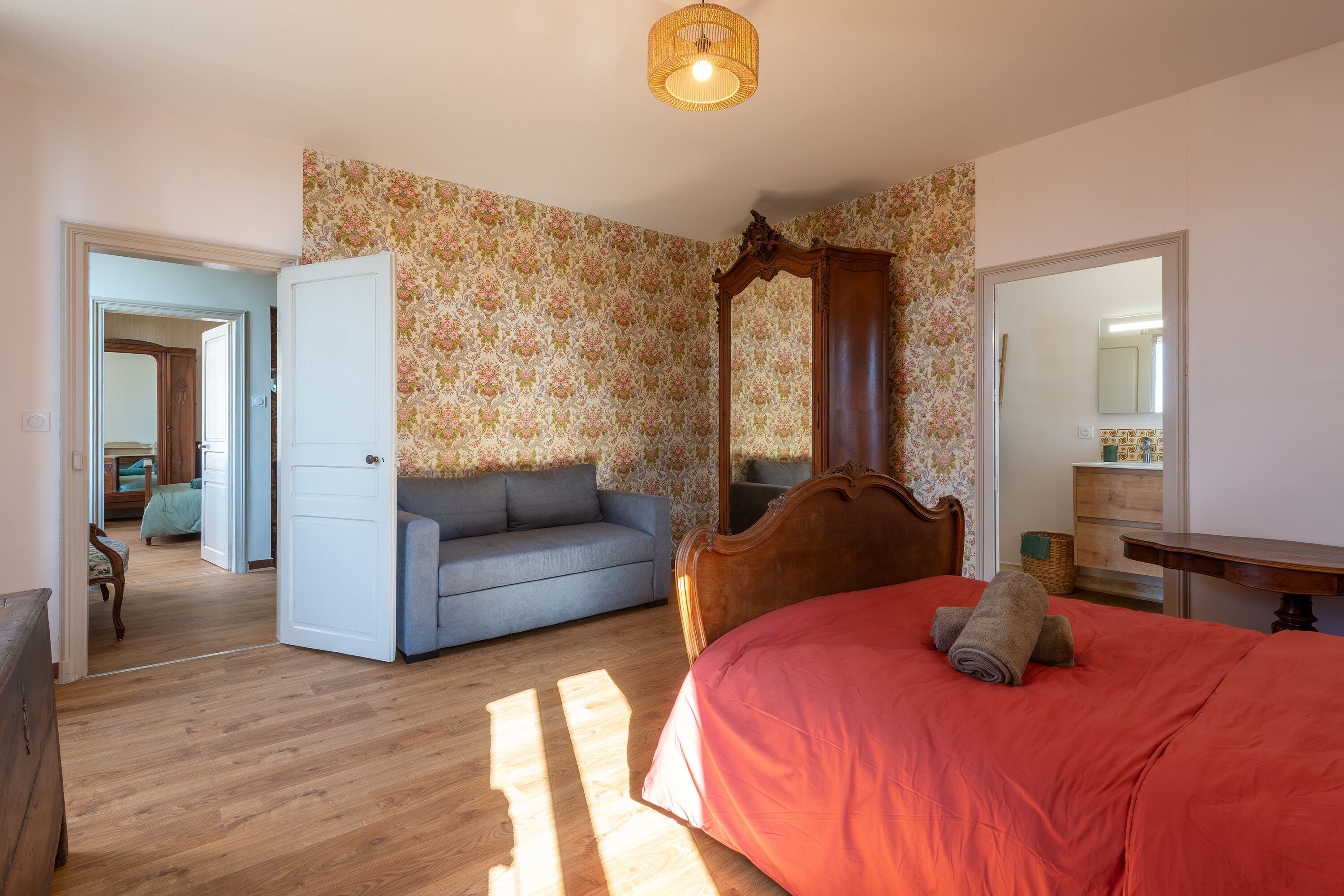 Un dormitorio con piso de madera, cama con dosel, sofá gris y papel tapiz floral.
