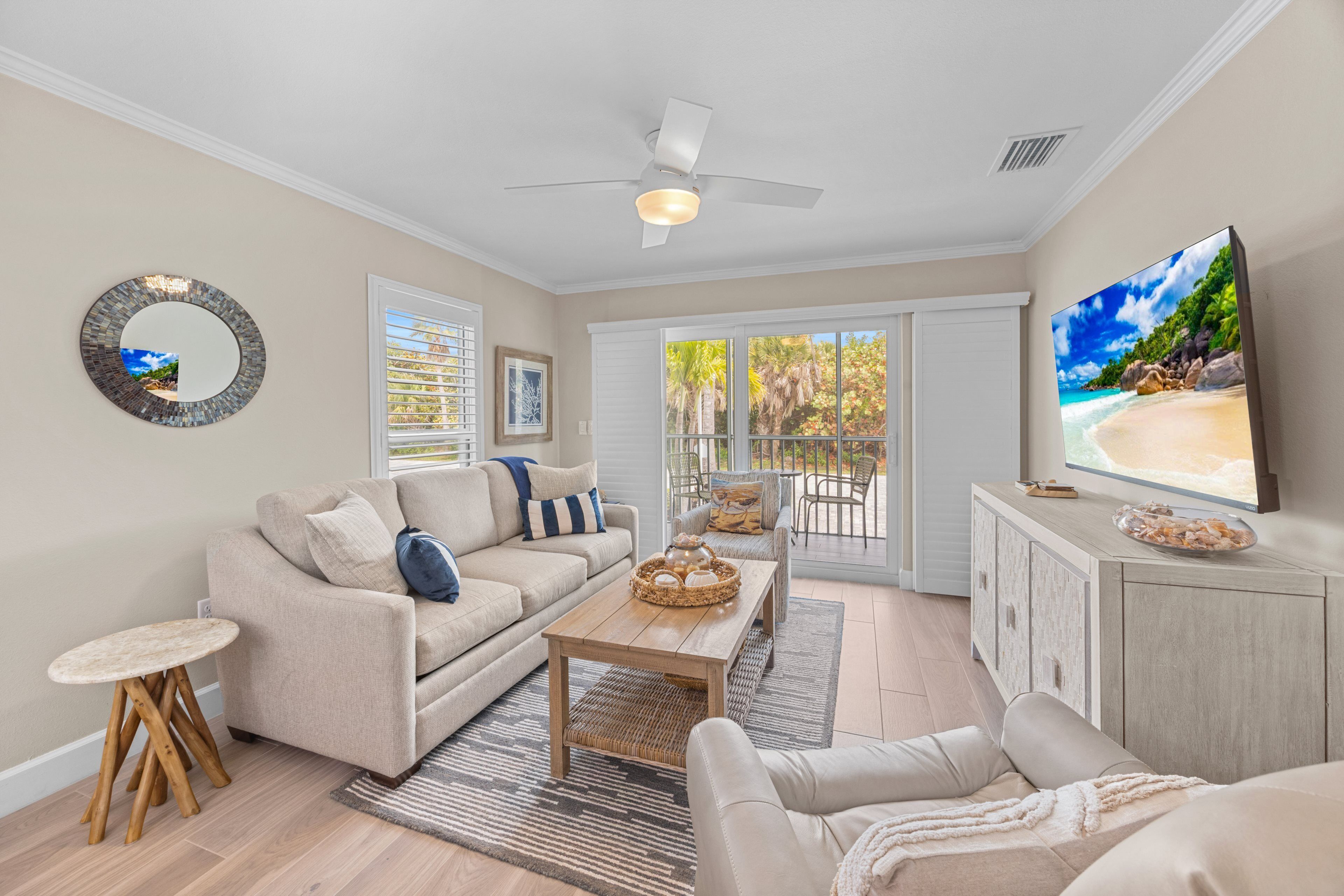 A living room with a sofa, coffee table, and TV. There is a balcony with a view of the outdoors.