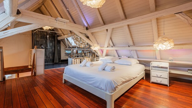 Una camera da letto con pavimento in legno, un letto con lenzuola bianche, un comodino e un soffitto a vista con travi.