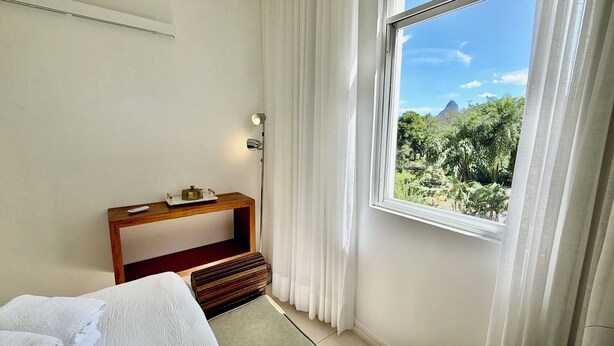 Quarto de hotel com cama, mesa de consola de madeira, mesa de cabeceira, luminária e janela com vista para vegetação e uma montanha.