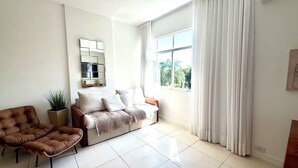 Uma sala de estar com um sofá, uma cadeira, uma planta em vaso e uma janela com cortinas.