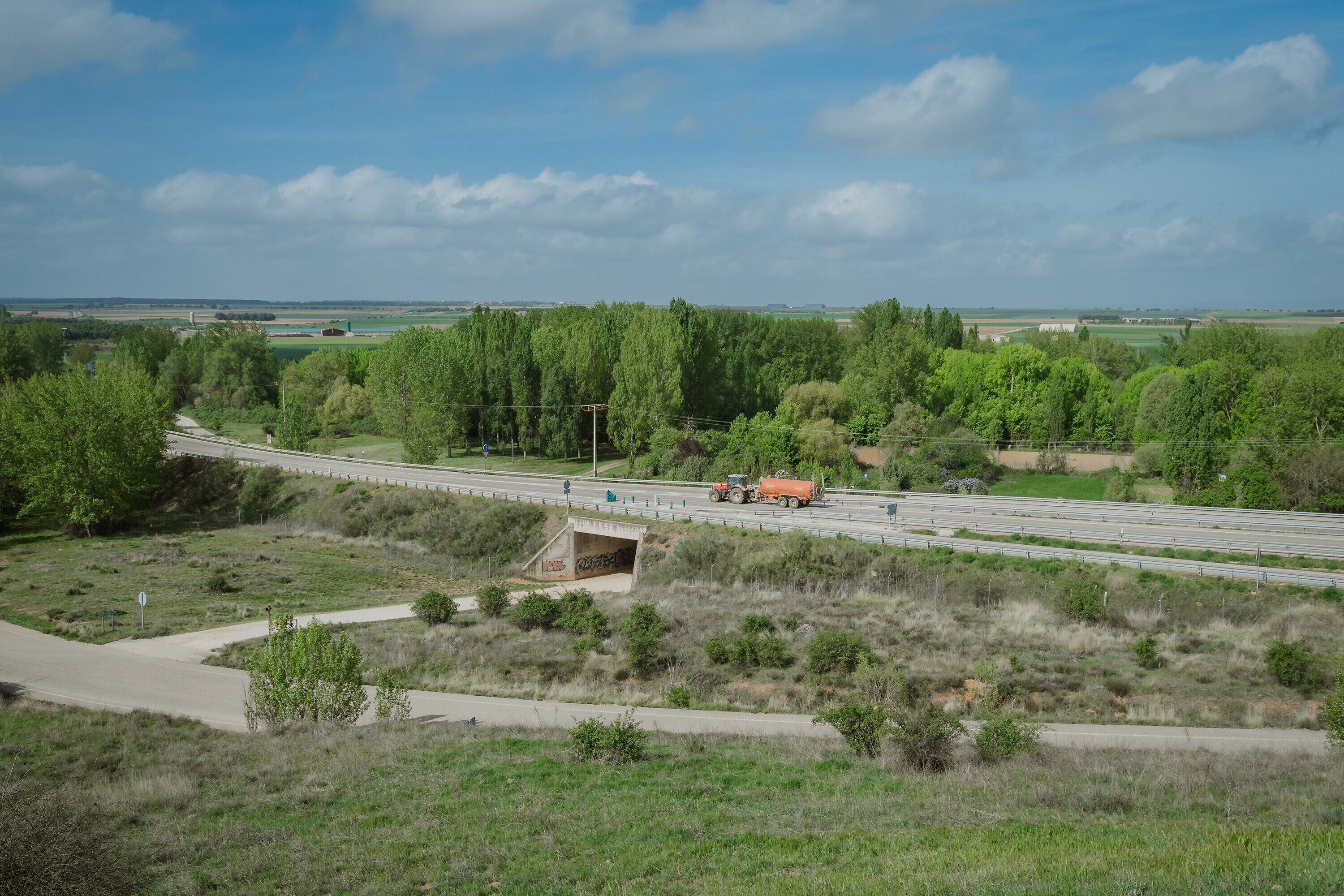 A highway with a bridge, greenery, and distant fields.