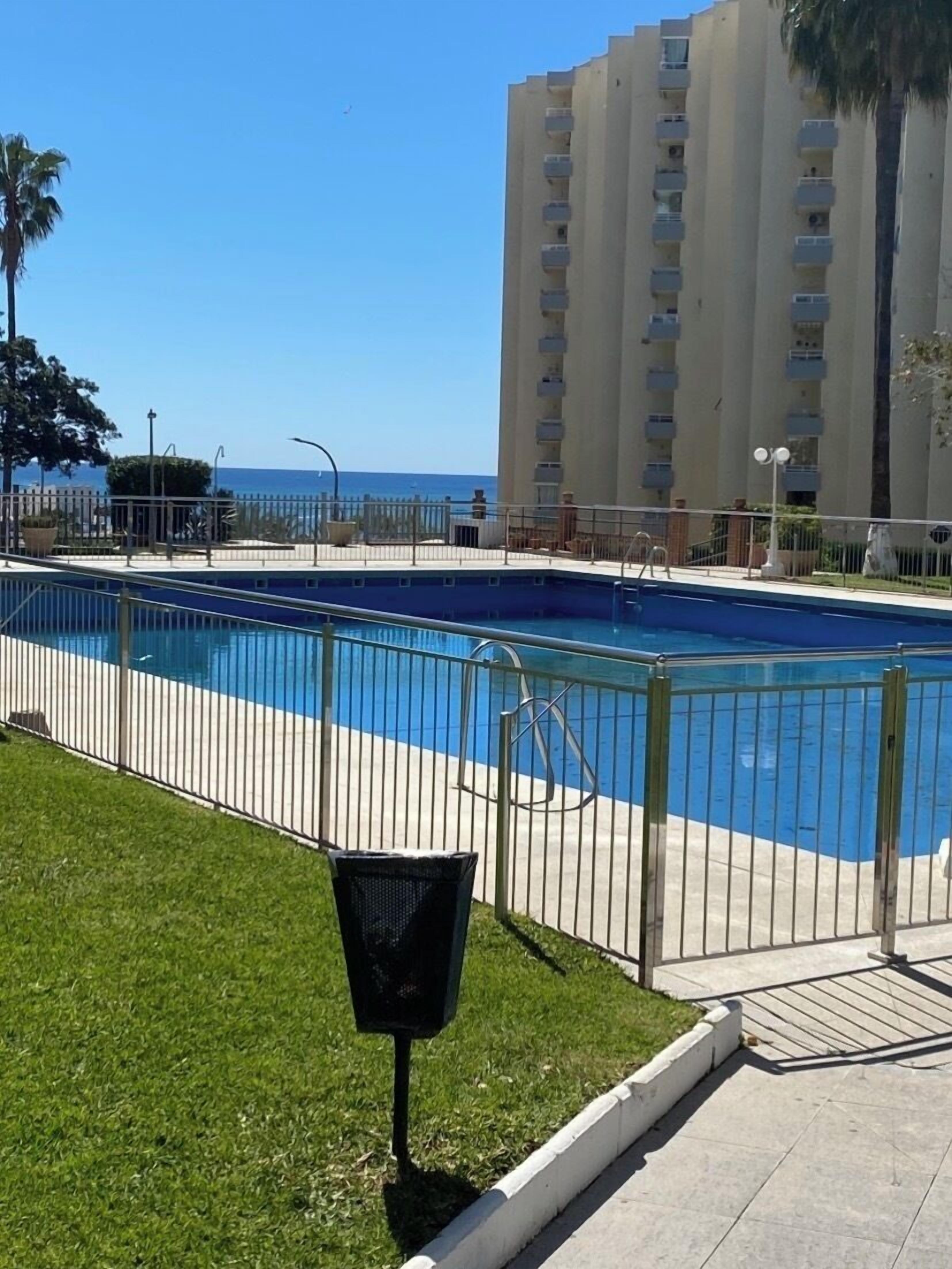 A swimming pool with a metal fence, a grassy area, and a tall building in the background.