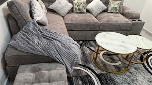 En modulsofa med mange puder, et gråt plaide og et rundt sofabord med gyldne ben.