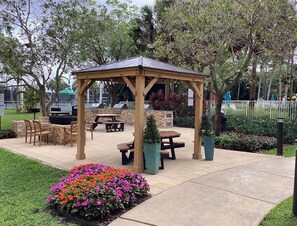 A gazebo with a roof, surrounded by outdoor seating, a barbecue grill, and a flower bed.