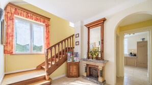 Eine Treppe führt zu einer Zwischenebene mit Kamin, im Hintergrund befindet sich ein Küchenbereich und ein Fenster mit Vorhängen.