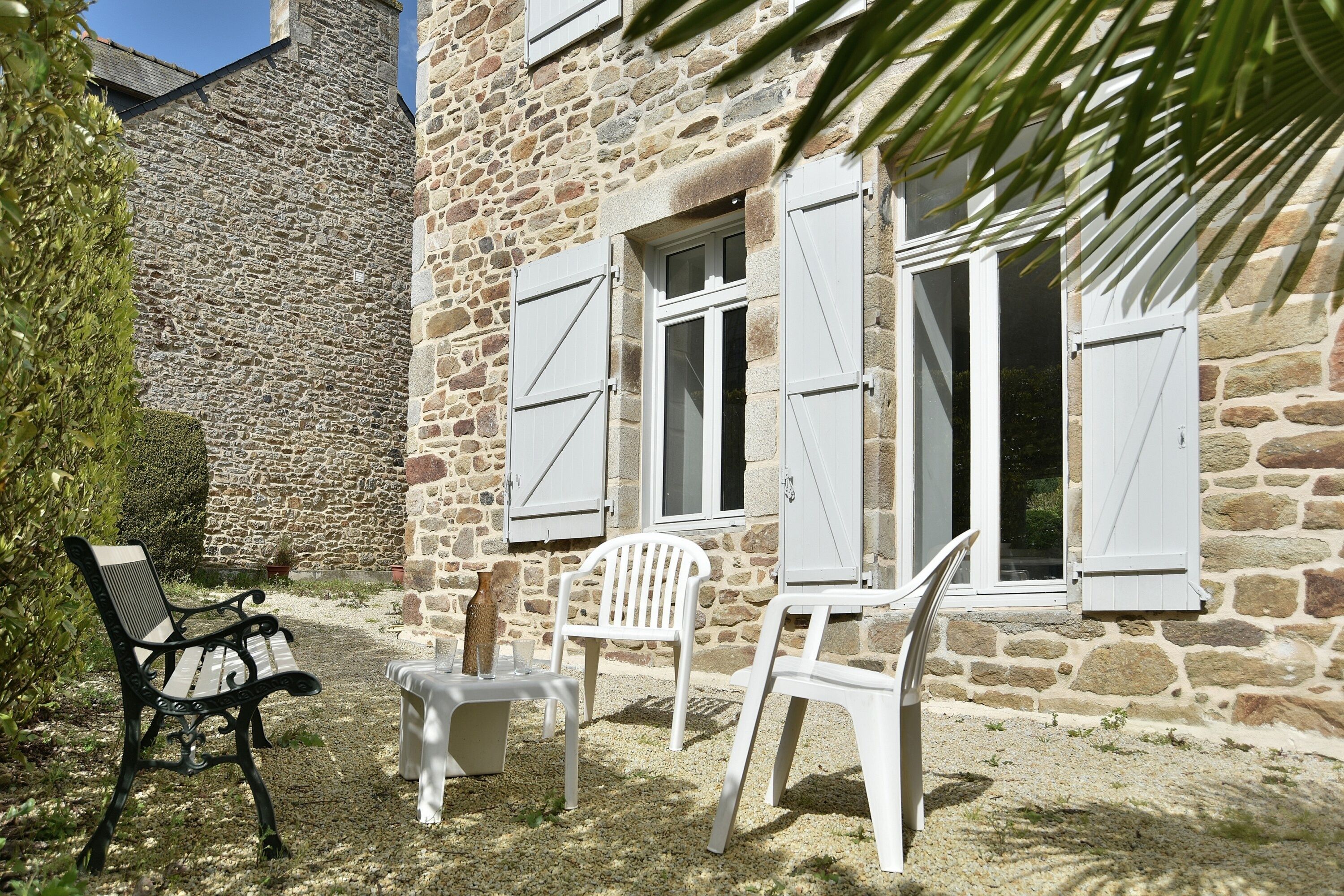 A stone building with white shutters, a bench, and two white plastic chairs.
