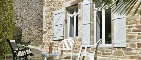 A stone building with white shutters, a bench, and two white plastic chairs.