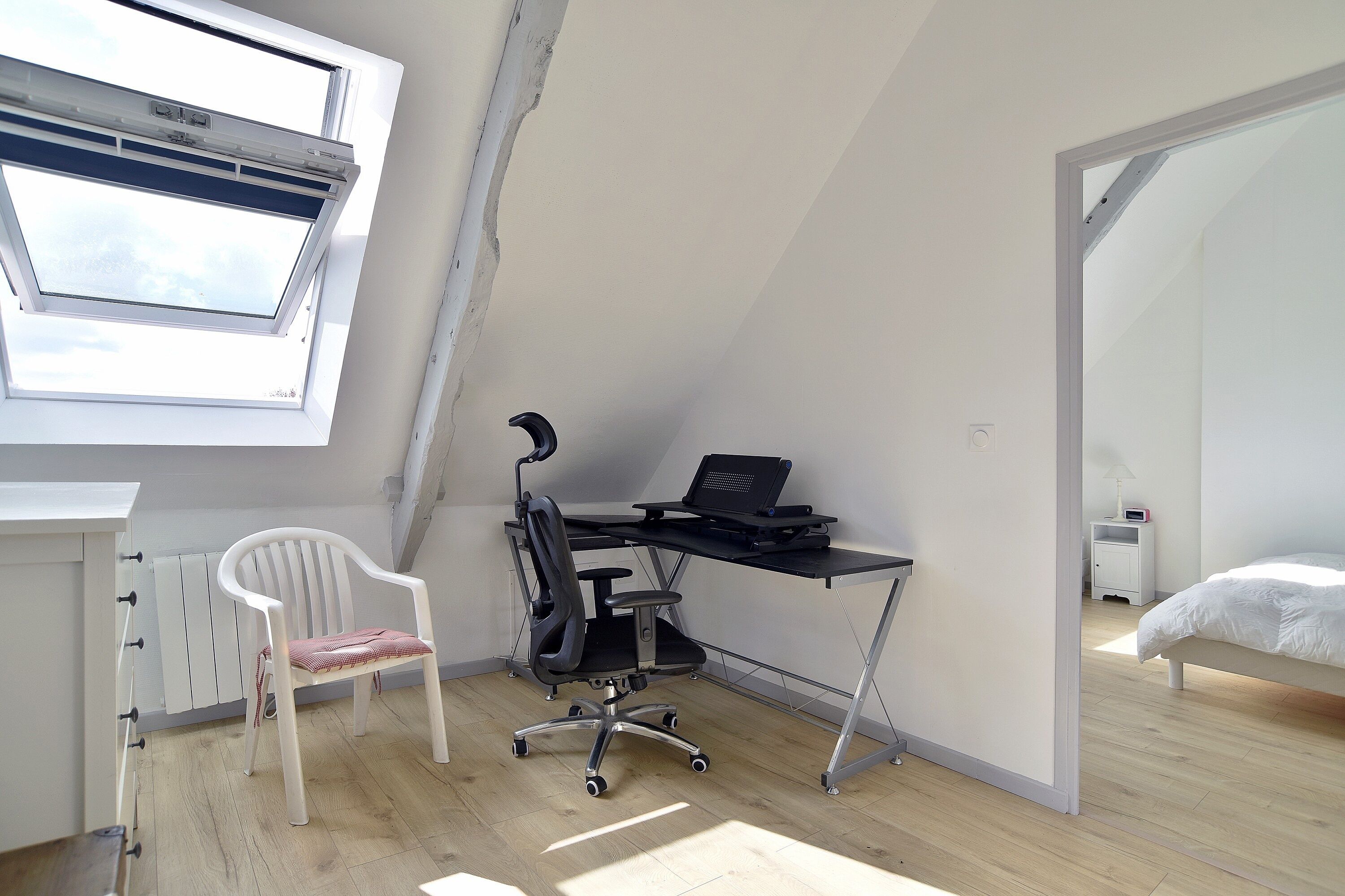 A room with a skylight, a desk with a computer, a white chair, and a bed.