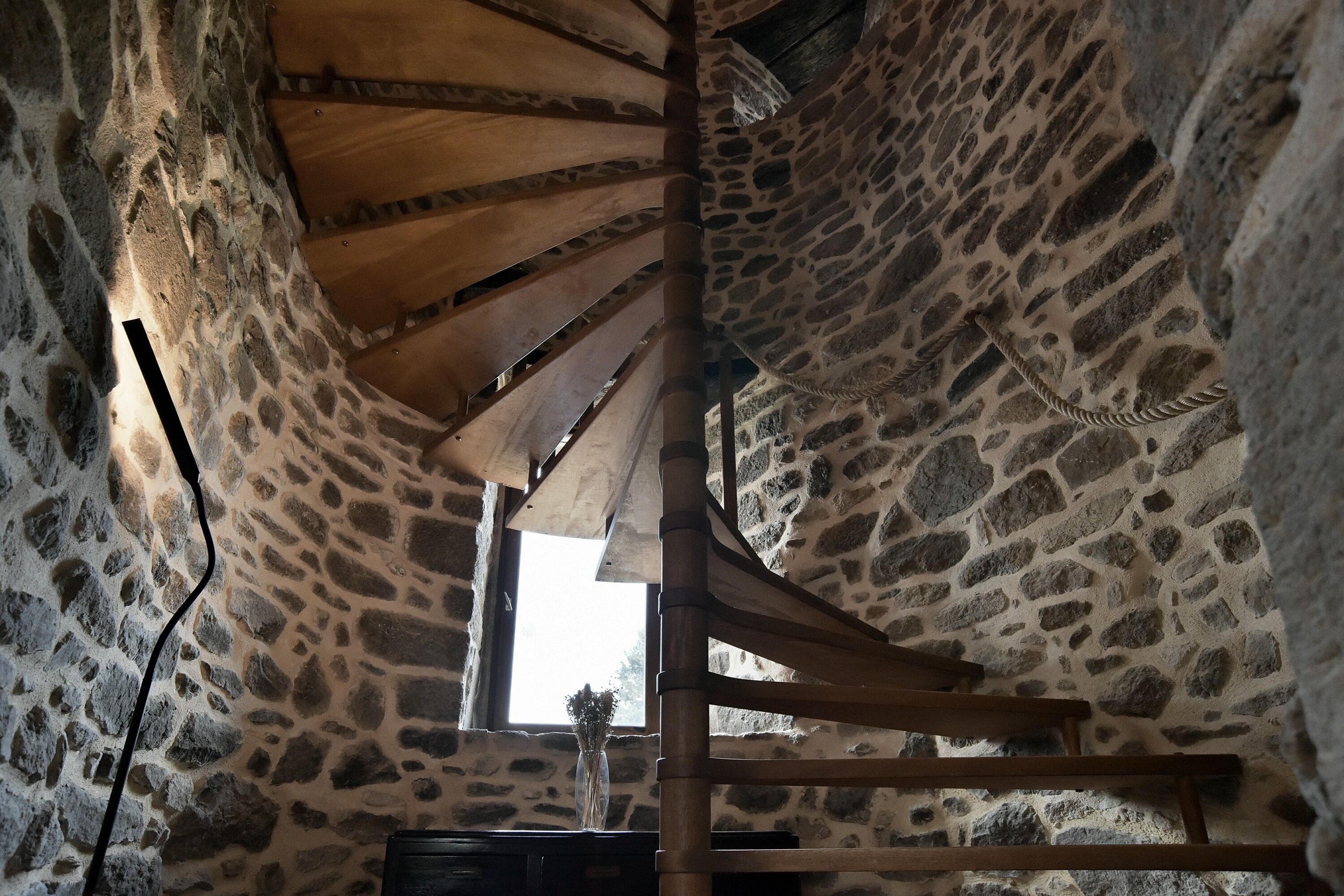 A stone-walled interior with a wooden spiral staircase, a window with a view, and a vase with flowers.
