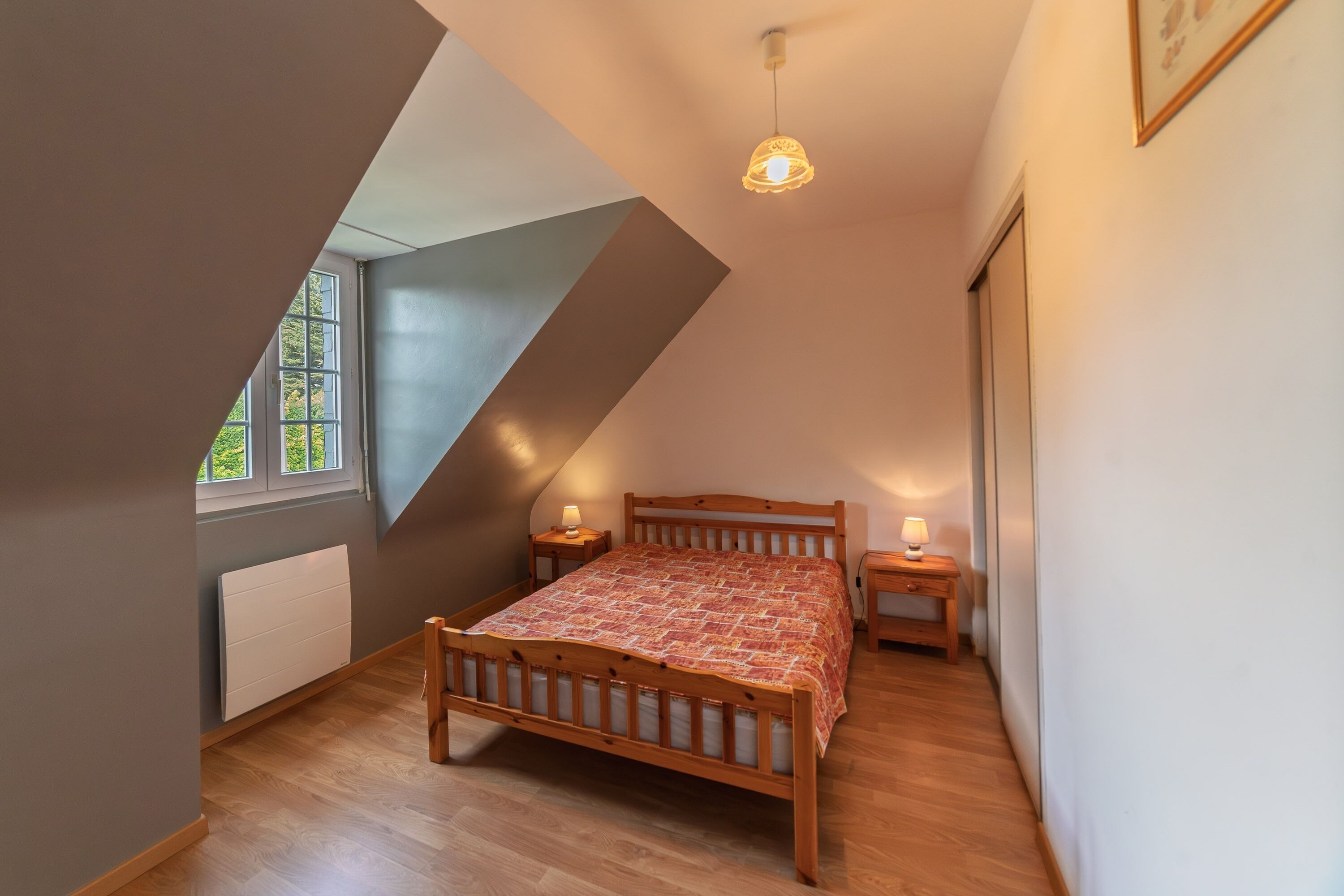 Un dormitorio con techo inclinado, una cama de madera, dos mesitas de noche con lámparas, un radiador y una ventana con vistas a la vegetación.