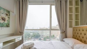 A hotel room with a large window, a bed with white linens, a headboard, and a bedside table. - Padi Suites Casa Grande, 2BR Apartment Mall Access (South Jakarta)