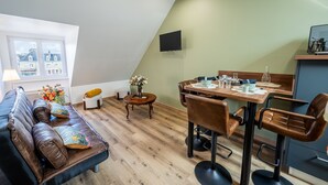 A modern living room with a sofa, dining table, and chairs, a flat-screen TV mounted on the wall, and a window with a view of buildings. - Le Tripot - A 15 min de la plage (Montebourg)