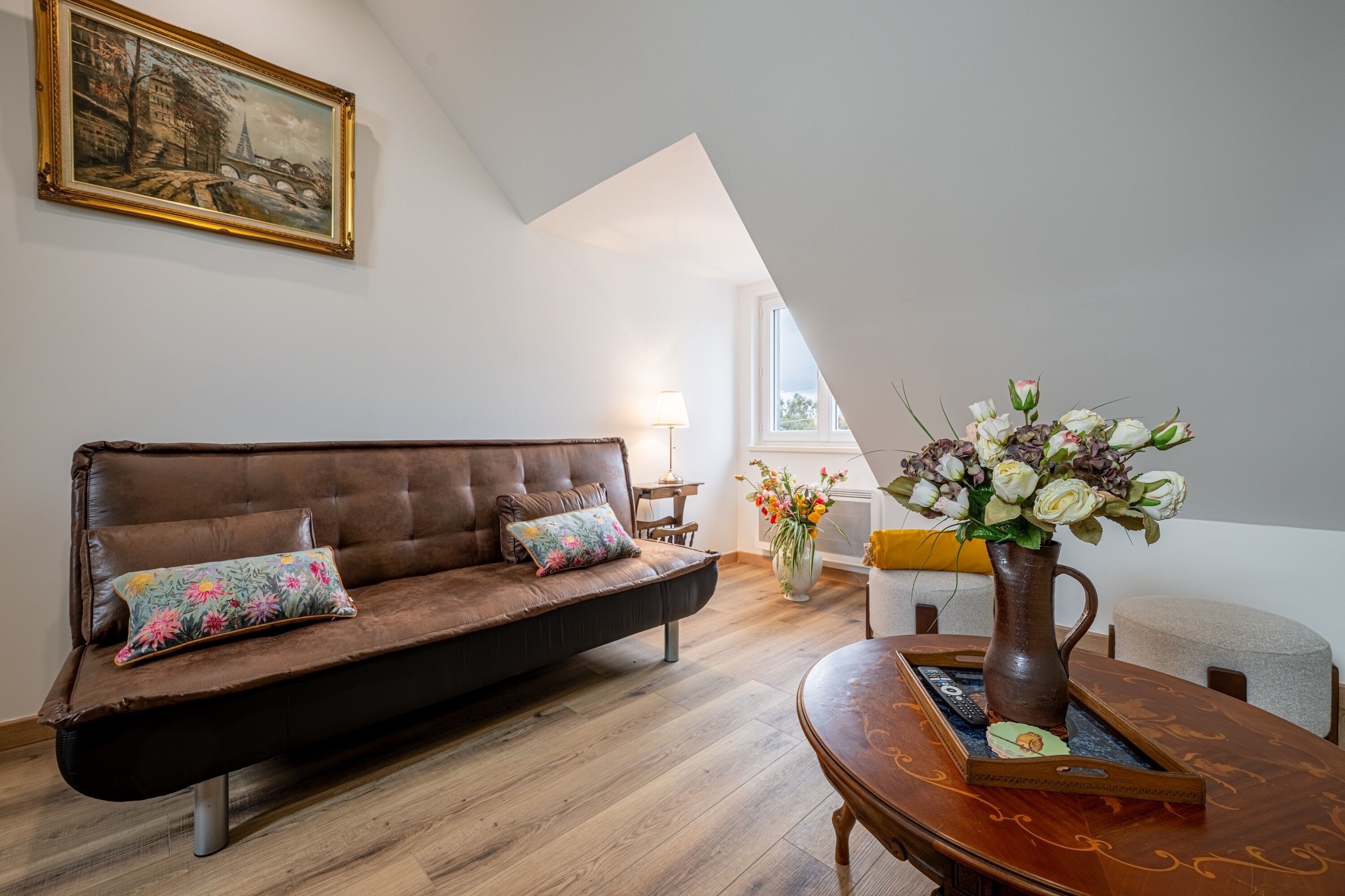 Un salon avec un canapé marron, une table basse en bois, un vase de fleurs, un tableau au mur et une fenêtre donnant sur un paysage.