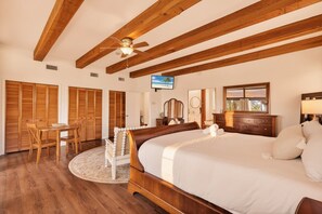 Une chambre avec un grand lit, des poutres apparentes en bois au plafond, un ventilateur de plafond, une télévision, un bureau avec des chaises et un tapis rond.