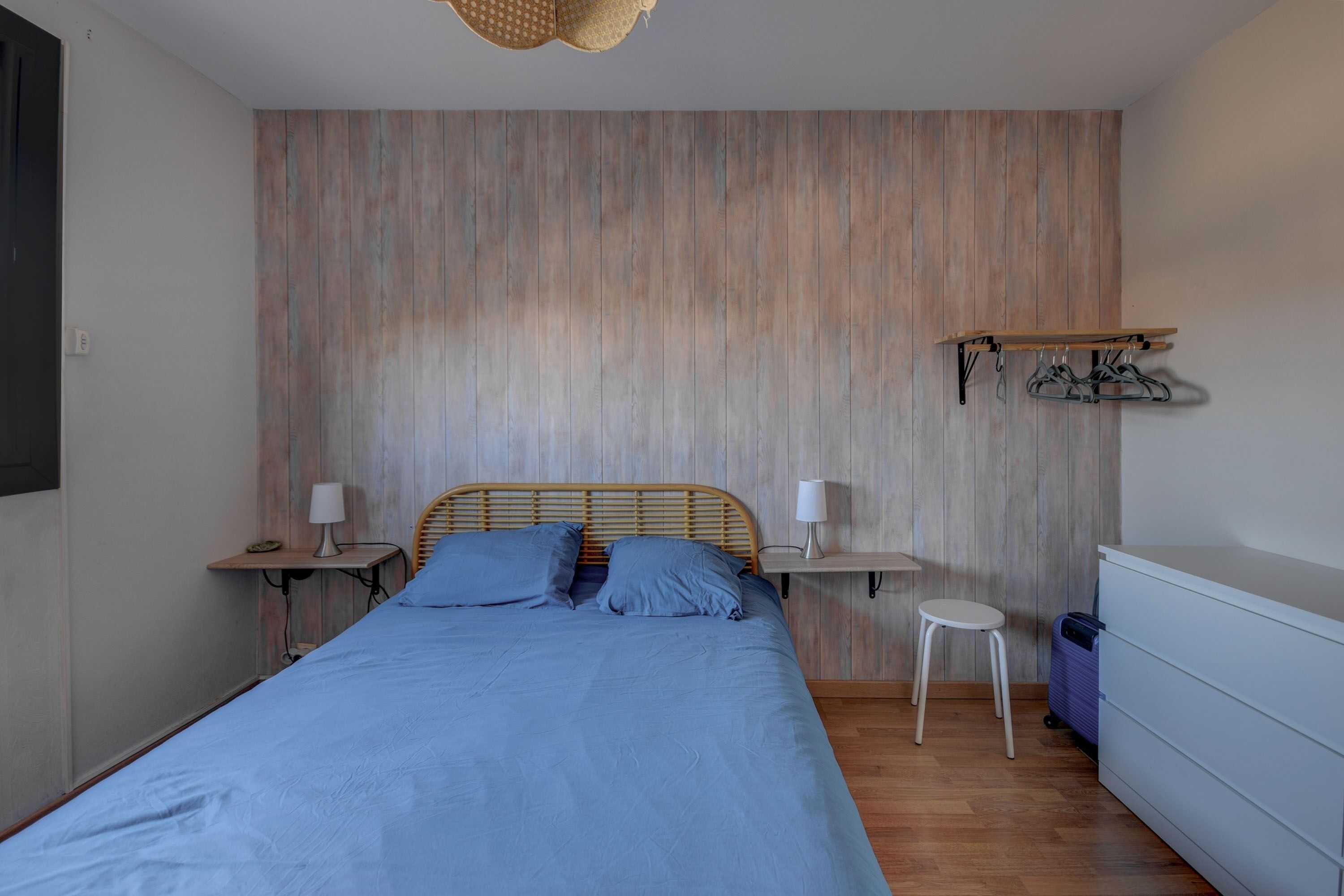 Une chambre avec un mur en bois, un lit avec une literie bleue, deux tables de chevet avec des lampes, un petit tabouret et une commode.