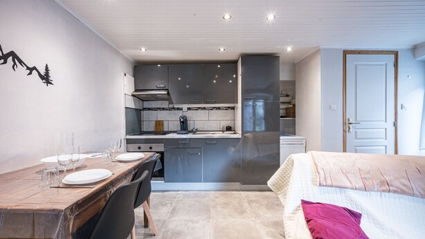 Une cuisine compacte dotée d’appareils électroménagers modernes, un coin repas avec une table dressée pour deux personnes, et un lit avec un seul oreiller.