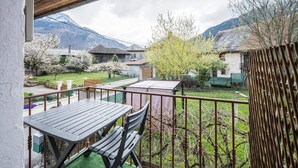 Un balcon avec une table et des chaises en bois, une vue sur un jardin arboré et une montagne en arrière-plan.