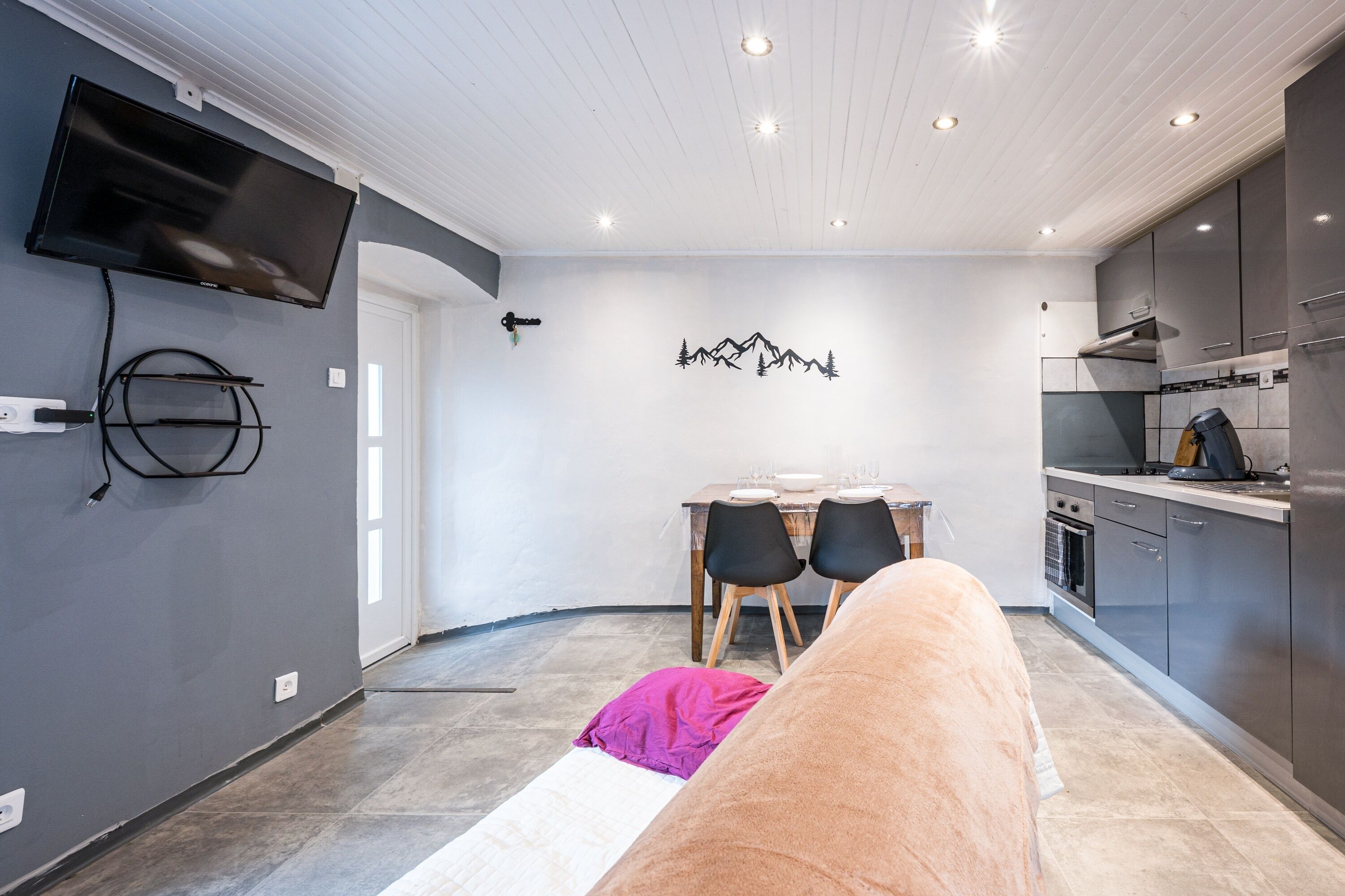A modern kitchen with a flat-screen TV, dining area with chairs, and a wall-mounted artwork.