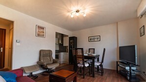Ein Wohnzimmer mit einer Couch, Sesseln, einem Esstisch, einem Fernseher und einer Küchenzeile.