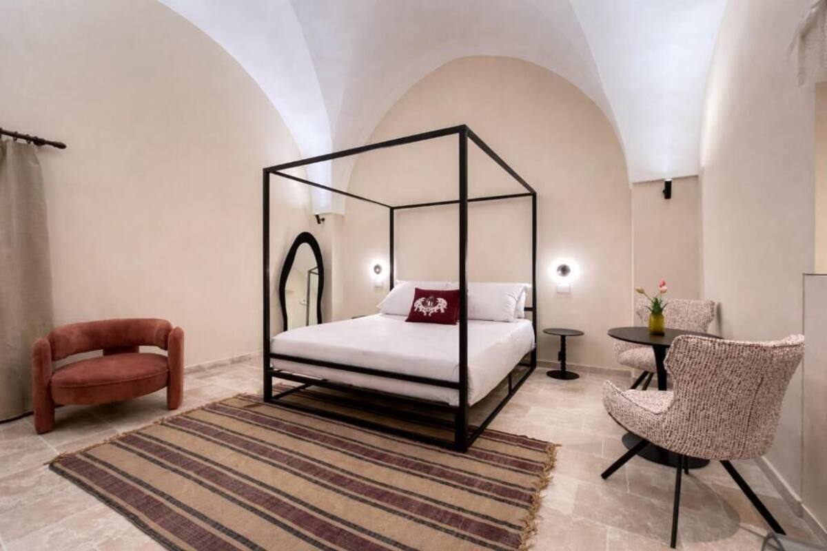 A bedroom with a four-poster bed, a red chair, a round table with a vase, and a small round table with a lamp.