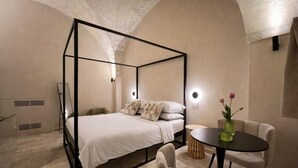 Una cama con dosel en una habitación con una mesa redonda y sillas, un jarrón con flores y una lámpara de pared.
