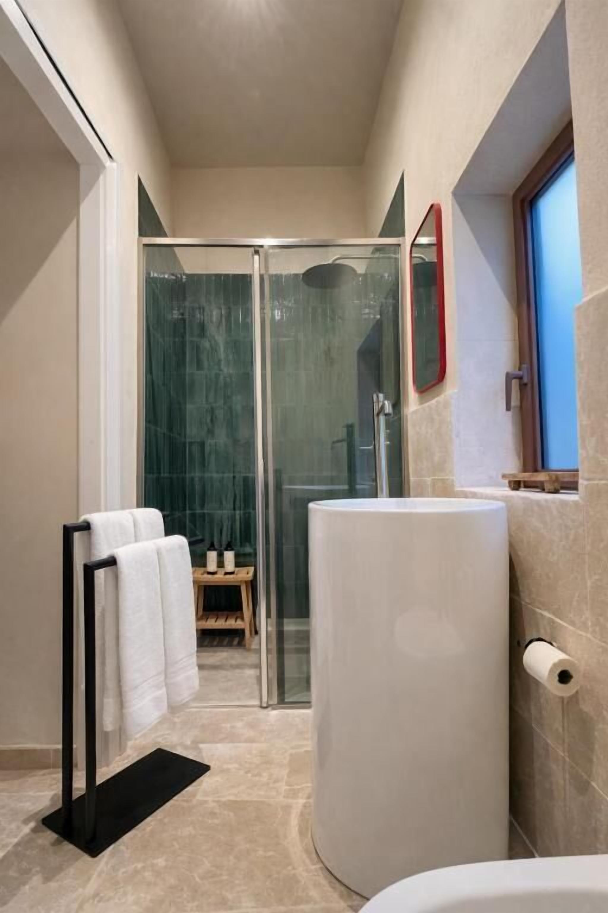 Ein modernes Badezimmer mit Duschkabine aus Glas, einem großen weißen Waschbecken und einem hölzernen Hocker mit Handtüchern.
