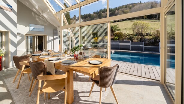 Un comedor con un porche de cristal que da a una piscina y un paisaje pintoresco.