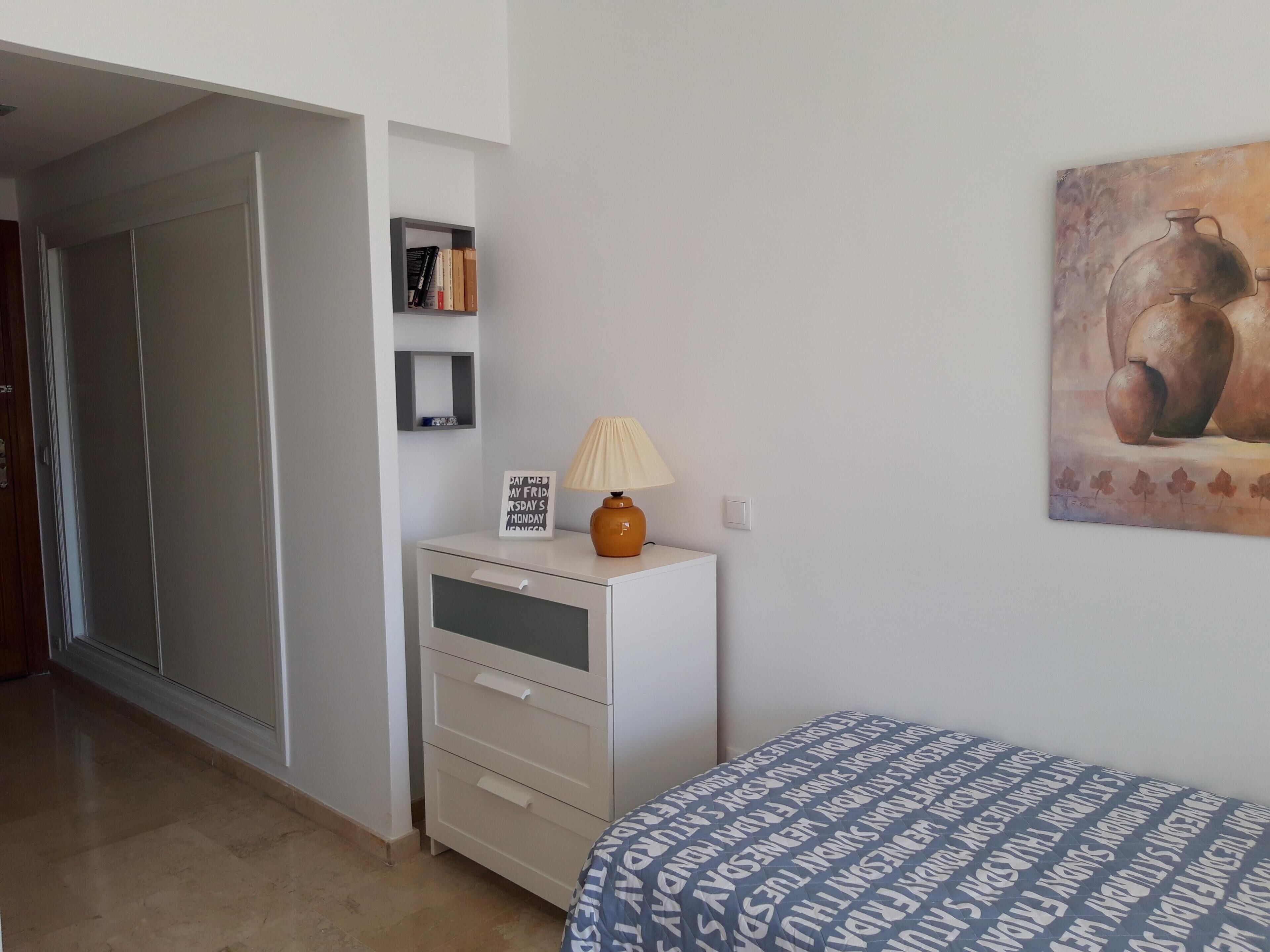 Ein Schlafzimmer mit einem Bett, einem Nachttisch, einer Lampe, einem an der Wand befestigten Regal mit Büchern, einer Tür und einem Gemälde.
