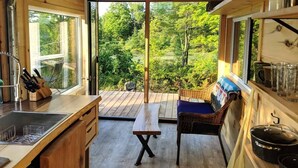 Interior de madera con un amplio ventanal corredero que da a una terraza, una zona de cocina con pileta y utensilios, y una silla de mimbre.