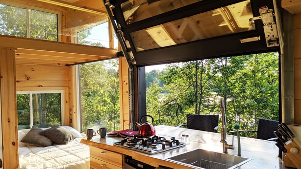 Una cabaña compacta y moderna con kitchenette, una cama y grandes ventanales con vistas al bosque.