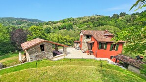 Une propriété située sur une colline, avec des bâtiments en pierre et aux façades rouges, une terrasse dallée et une végétation luxuriante tout autour.