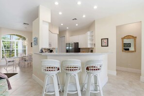 A modern kitchen with white cabinetry, a central island with bar stools, and a dining area with wicker chairs. - Naples Vineyards Living | Private Pool + Spa (Naples)