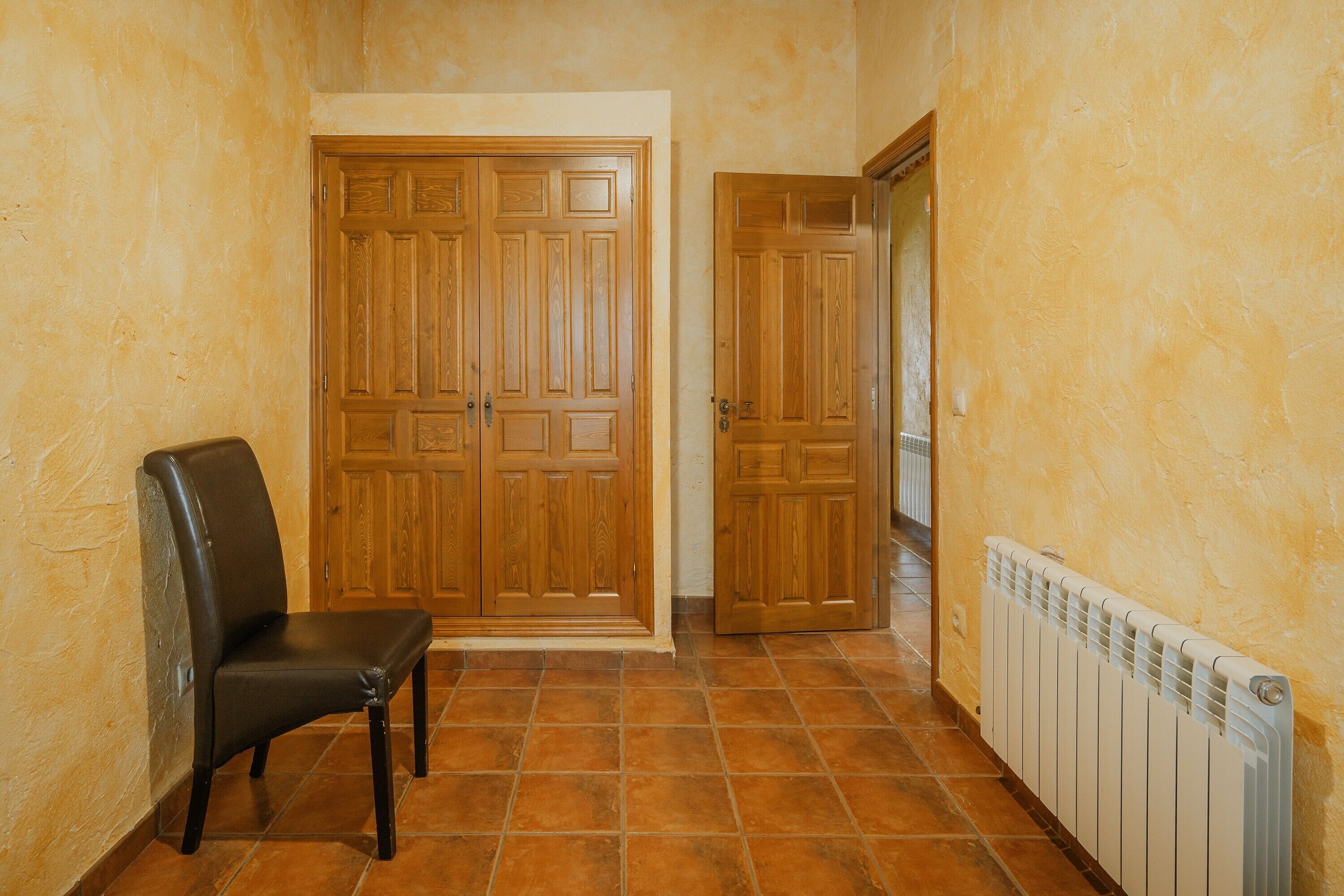 Un couloir avec une porte double en bois, un radiateur et un fauteuil en cuir noir.