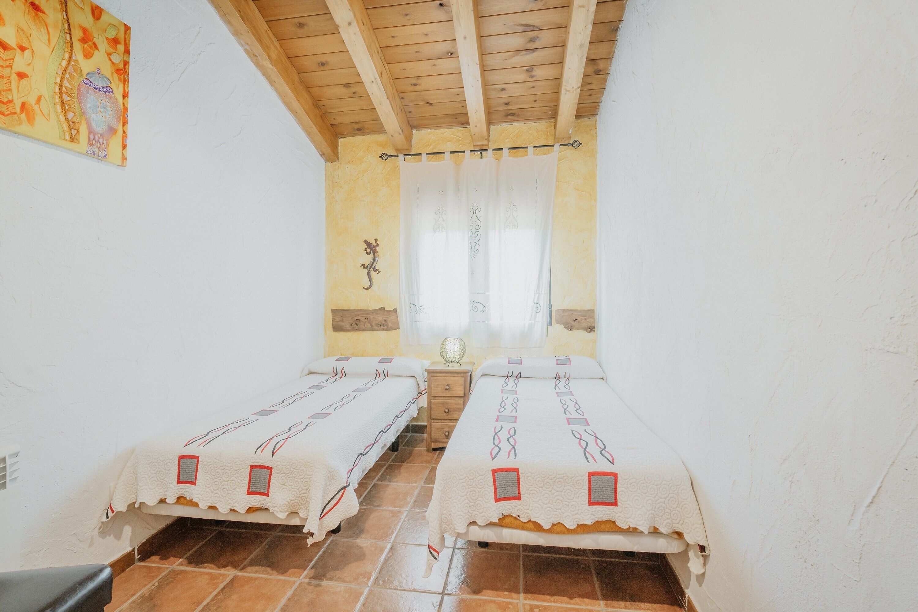 A room with two beds, wooden ceiling beams, a wall-mounted heater, and a window with curtains.