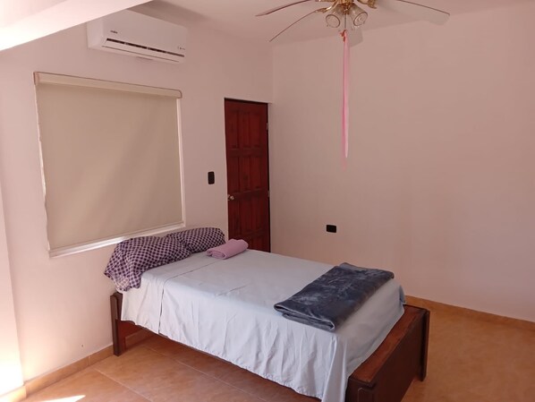 Un dormitorio con cama, ventana con persiana, unidad de aire acondicionado, puerta de madera y ventilador de techo.