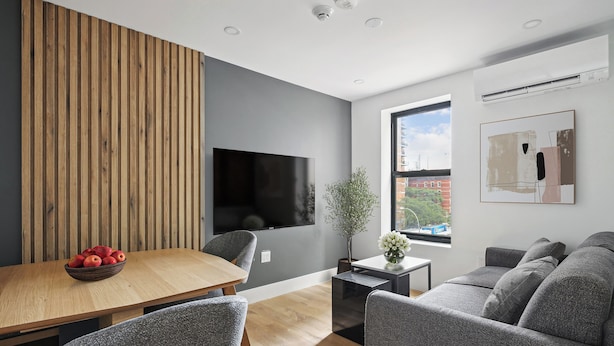 Ein modernes Wohnzimmer mit Wandverkleidung aus Holz, einem Flachbildfernseher, einem Esstisch mit Obstschale, grauen Polstermöbeln und einem Fenster mit Blick auf Gebäude.