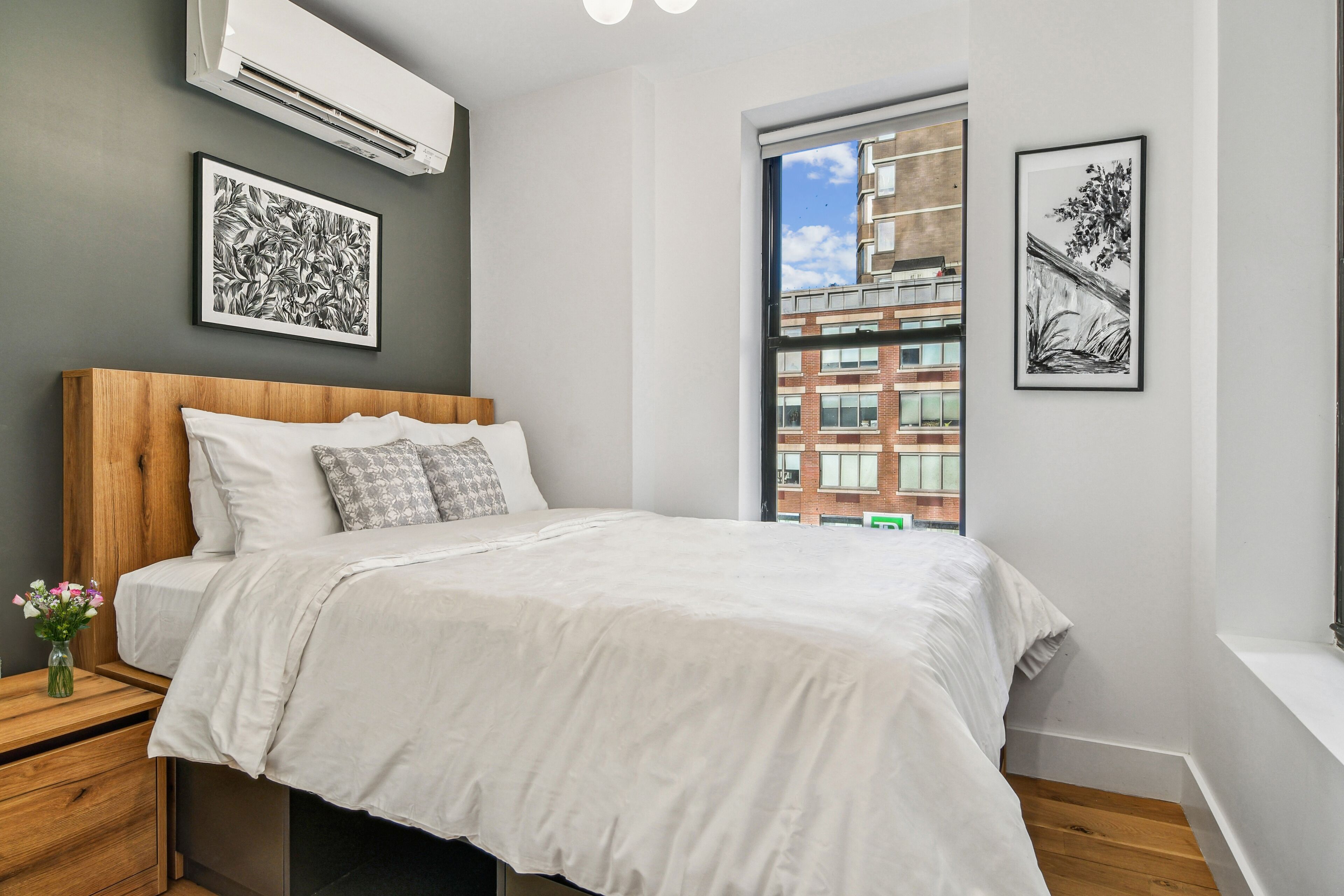 A modern bedroom with a wooden headboard, a large bed, a nightstand with a vase, and two framed artworks on the walls.